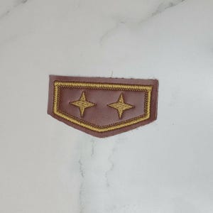 May include: A brown, shield-shaped patch featuring a gold embroidered border and two gold embroidered four-pointed star icons centered within the shape. The patch has a smooth, matte finish and a distinct, pointed bottom edge design.