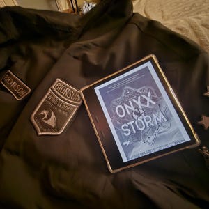 May include: A dark green jacket with two embroidered patches, one reading "RIORSON" and the other "RIORSON WING LEADER FIFTH WING". A digital tablet shows the book cover "ONYX STORM".