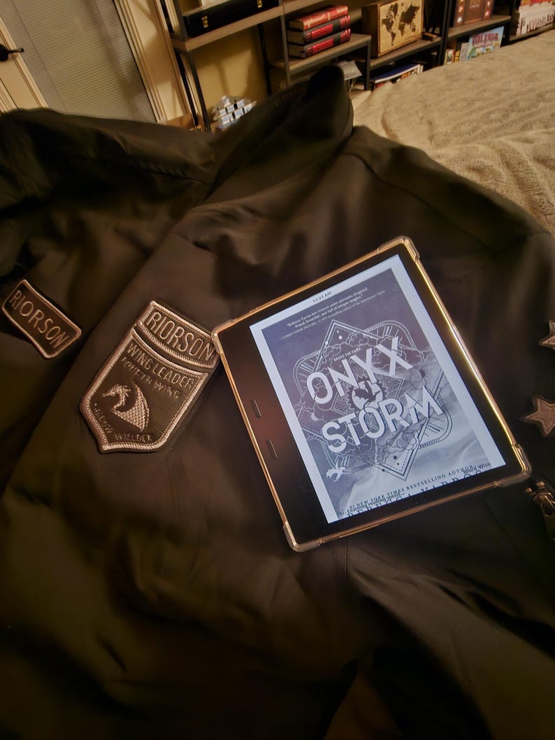 May include: A dark green jacket with two embroidered patches, one reading "RIORSON" and the other "RIORSON WING LEADER FIFTH WING". A digital tablet shows the book cover "ONYX STORM".