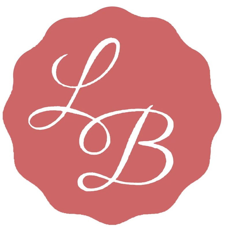 May include: A logo featuring the white cursive letters "L" and "B" on a scalloped, dusty rose-colored background. The letters are intertwined in a flowing, elegant script.