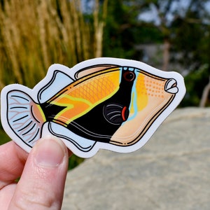 May include: A colorful sticker featuring a stylized fish with a black, orange, and yellow body. The fish has a white outline and a light blue fin. The sticker is held by a hand against a natural background.