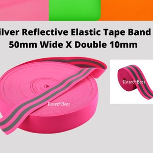 Peut inclure: Bande &eacute;lastique rose avec deux bandes r&eacute;fl&eacute;chissantes argent&eacute;es. La bande mesure 50 mm de large et les bandes r&eacute;fl&eacute;chissantes 10 mm de large. La bande est enroul&eacute;e et la marque "Rosec&Bees" est visible sur la bande.