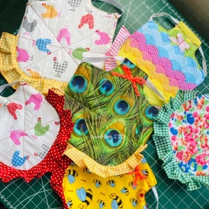 May include: A collection of colorful, handmade pot holders with various patterns. Designs include chickens, peacock feathers, bees, and floral prints. Each pot holder has a ruffled edge and a loop for hanging.
