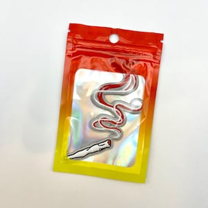 May include: Embroidered patch depicting a stylized joint with red smoke. The patch is in a clear, iridescent pouch with a red and yellow gradient border. The patch is white, grey, and red.