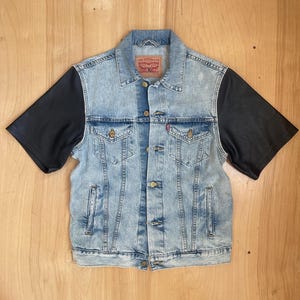 May include: A light-wash denim jacket with black short sleeves. The jacket features a classic collar, button-up front, and two chest pockets. A red Levi Strauss & Co. tag is visible on the back of the collar.