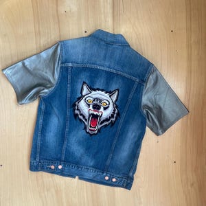 Puede incluir: Chaqueta vaquera azul con mangas cortas plateadas. La parte trasera de la chaqueta presenta una gran cabeza de lobo bordada con dientes afilados. La chaqueta tiene una parte delantera abotonada y un cuello.