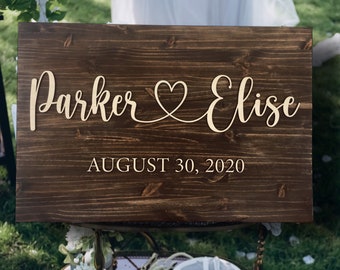 May include: A rustic wooden wedding sign with the names "Parker & Elise" and the date "August 30, 2020" in white lettering. The sign is on a vintage cart decorated with white flowers.
