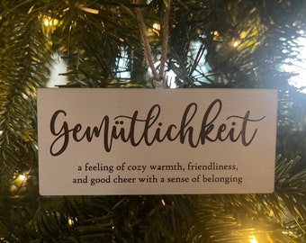 May include: A wooden Christmas ornament with the German word "Gemütlichkeit" and the definition: "a feeling of cozy warmth, friendliness, and good cheer with a sense of belonging."