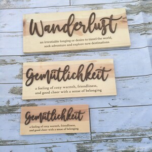 May include: Three wooden signs with the words "Wanderlust", "Gem&uuml;tlichkeit", and "Gem&uuml;tlichkeit" written in brown lettering. The signs are on a light wood background. The definition of each word is written below the word.