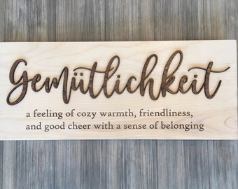 May include: A wooden sign with the German word "Gemütlichkeit" carved into it. The word is defined as "a feeling of cozy warmth, friendliness, and good cheer with a sense of belonging."