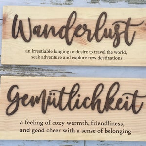 May include: Two wooden signs with the German words "Wanderlust" and "Gem&uuml;tlichkeit" and their definitions. The signs are made of light wood with dark brown lettering.
