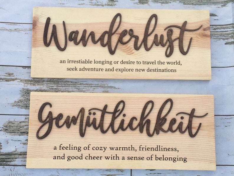 May include: Two wooden signs with the German words "Wanderlust" and "Gem&uuml;tlichkeit" and their definitions. The signs are made of light wood with dark brown lettering.