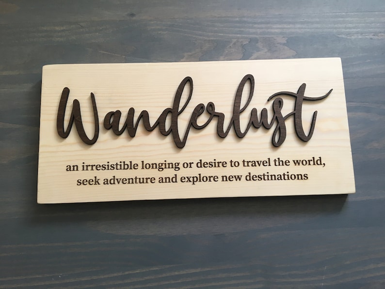 May include: Wooden sign with the word "Wanderlust" in a decorative font. The sign also includes the definition of wanderlust: "an irresistible longing or desire to travel the world, seek adventure and explore new destinations."