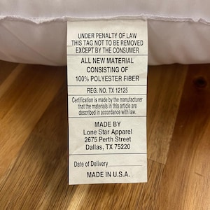 May include: A white pillow with a tag attached. The tag includes text that reads "UNDER PENALTY OF LAW THIS TAG NOT TO BE REMOVED EXCEPT BY THE CONSUMER" and "MADE IN U.S.A."