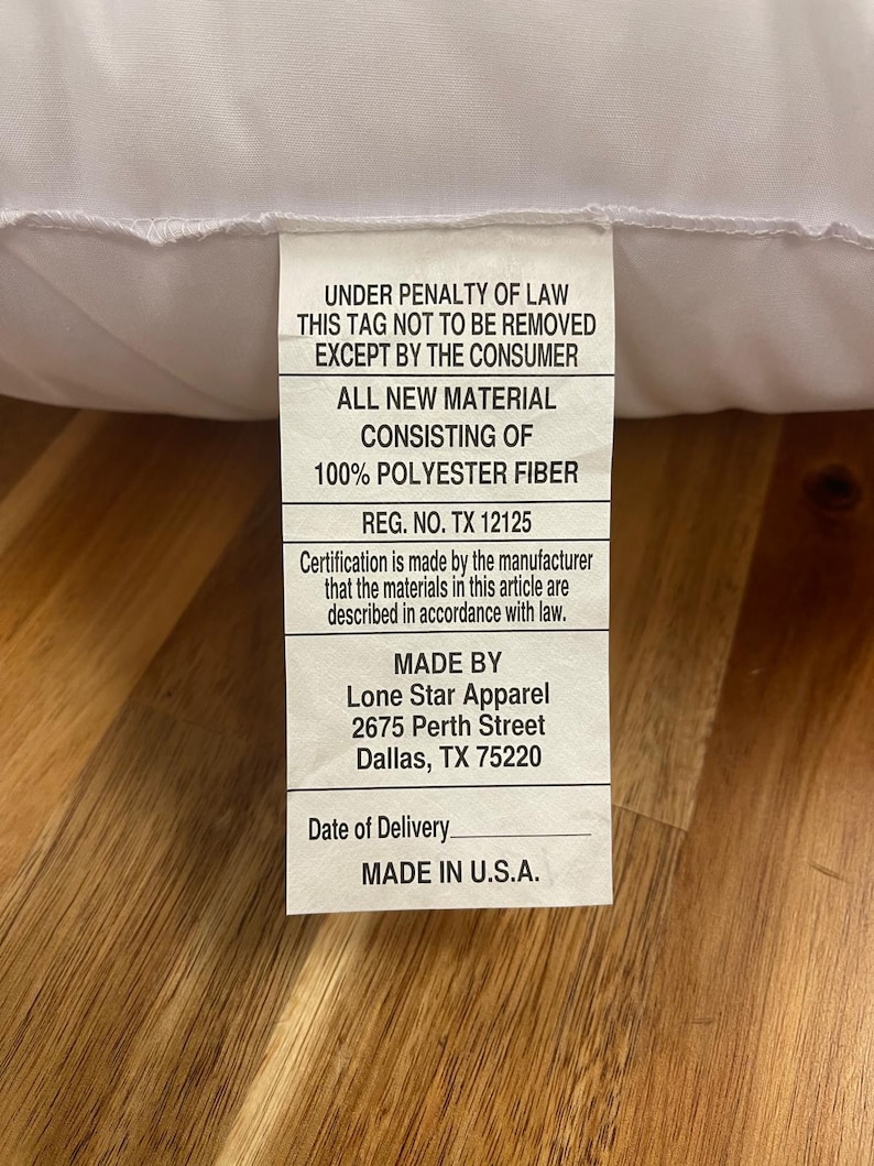 May include: A white pillow with a tag attached. The tag includes text that reads "UNDER PENALTY OF LAW THIS TAG NOT TO BE REMOVED EXCEPT BY THE CONSUMER" and "MADE IN U.S.A."
