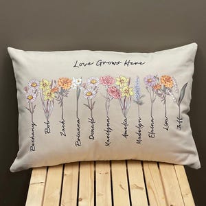 May include: Rectangular beige pillow with a floral design and the text "Love Grows Here." The pillow features various flowers in shades of yellow, pink, orange, and white, with names written below each flower.