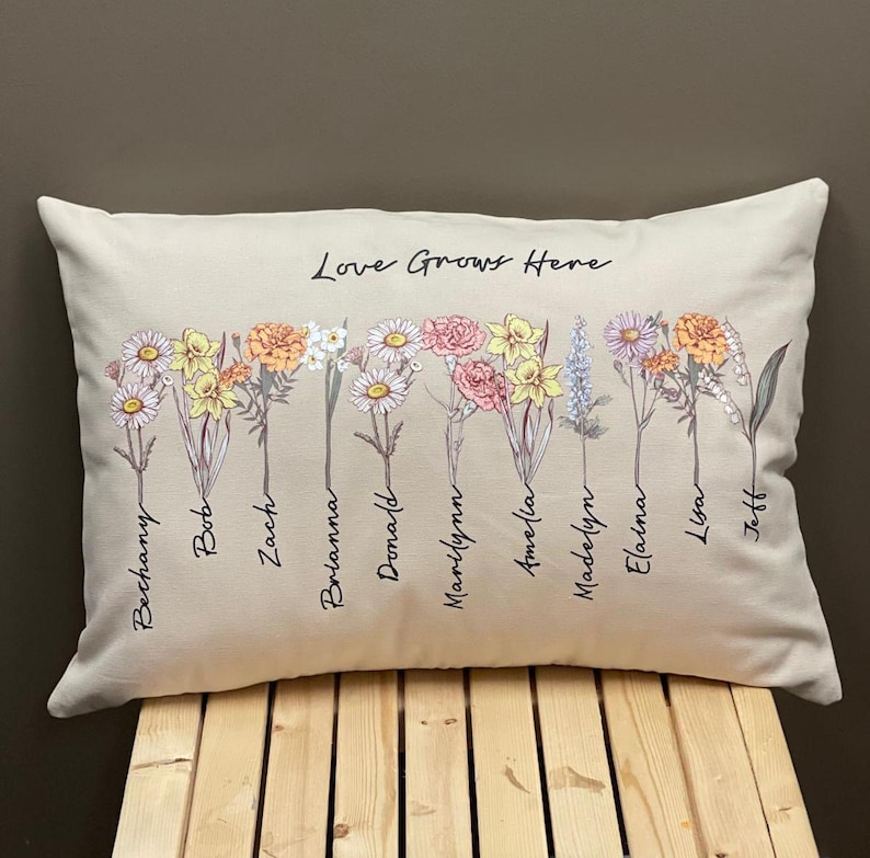 May include: Rectangular beige pillow with a floral design and the text "Love Grows Here." The pillow features various flowers in shades of yellow, pink, orange, and white, with names written below each flower.