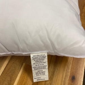 May include: A white pillow with a tag attached. The tag includes text that reads "UNDER PENALTY OF LAW THIS TAG NOT TO BE REMOVED EXCEPT BY THE CONSUMER ALL NEW MATERIAL CONSISTING OF 100% POLYESTER FIBER REG. NO. TX 12125 MADE IN U.S.A."