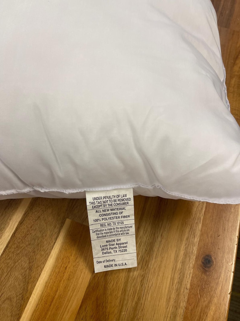 May include: A white pillow with a tag attached. The tag includes text that reads "UNDER PENALTY OF LAW THIS TAG NOT TO BE REMOVED EXCEPT BY THE CONSUMER ALL NEW MATERIAL CONSISTING OF 100% POLYESTER FIBER REG. NO. TX 12125 MADE IN U.S.A."