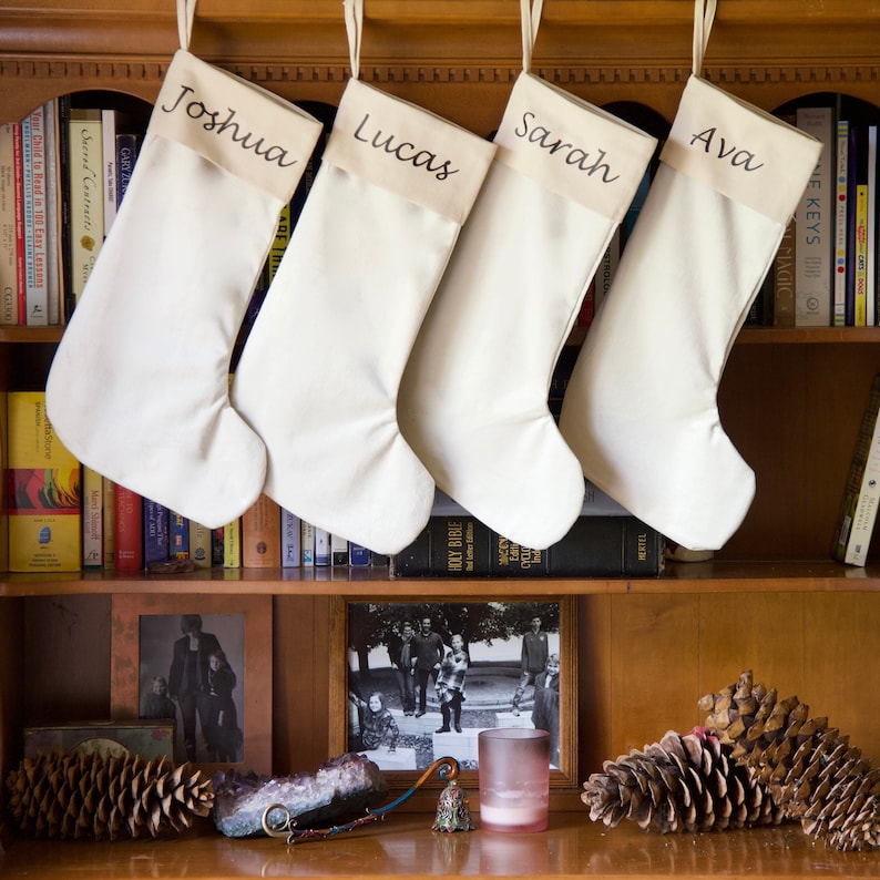 May include: Four white canvas stockings with names embroidered on them, "Joshua", "Lucas", "Sarah", and "Ava", hanging on a wooden shelf with books and decorations.