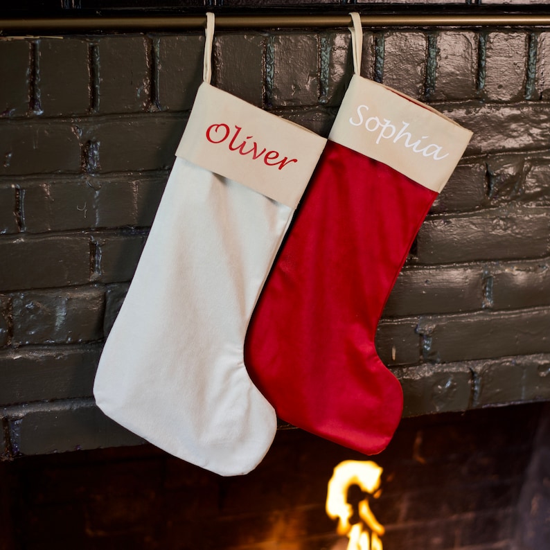 May include: Two Christmas stockings hanging on a fireplace mantel. The stocking on the left is white with the name "Oliver" embroidered on it. The stocking on the right is red with the name "Sophia" embroidered on it.