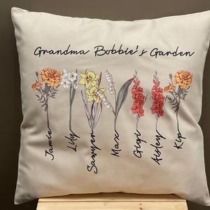 May include: Beige square throw pillow with the text "Grandma Bobbie's Garden" and floral illustrations. The pillow features various flowers with names like Jamie, Lily, and Sawyer. The flowers are in shades of yellow, orange, and red.