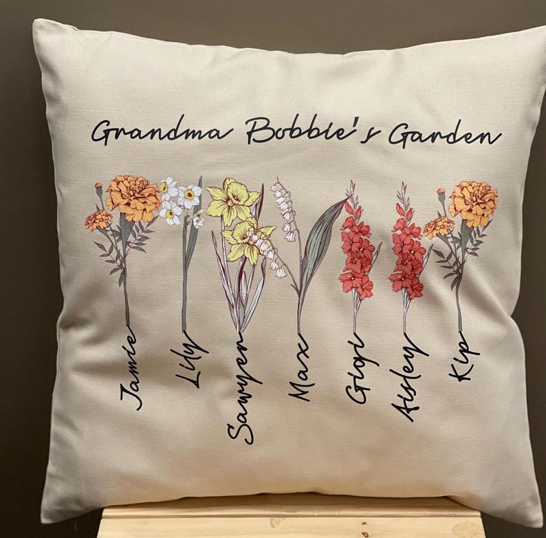 May include: Beige square throw pillow with the text "Grandma Bobbie's Garden" and floral illustrations. The pillow features various flowers with names like Jamie, Lily, and Sawyer. The flowers are in shades of yellow, orange, and red.