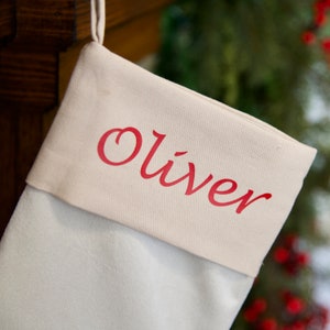 May include: A white canvas stocking with a red felt cuff. The cuff has the name "Oliver" written in red cursive.