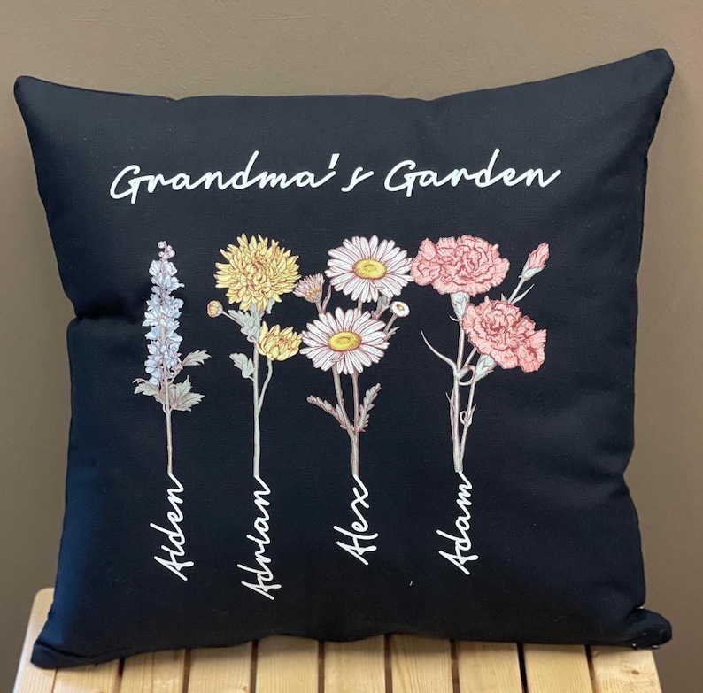 May include: Black decorative pillow with the text "Grandma's Garden" in white script. The pillow features four floral illustrations with names below each flower: Aiden, Adrian, Alex, and Adam. The flowers are in shades of white, yellow, and pink.