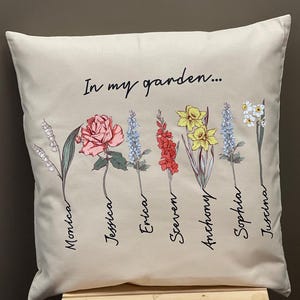May include: Beige square pillow with the text "In my garden..." and floral illustrations. The flowers include a rose, daffodils, and other blooms, each with a name below. The names are Monica, Jessica, Erica, Steven, Anthony, Sophia, and Justina.