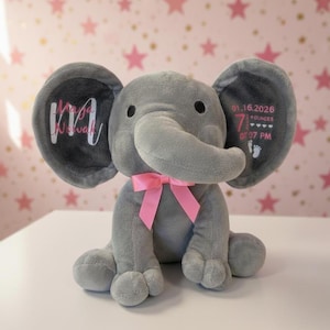 May include: A gray plush elephant toy with a pink bow, personalized with the name "Maja Nowak" on one ear and birth details on the other. The birth details include the date, time, and weight in ounces. The background features a pink and gold star pattern.