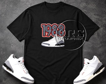 Peut inclure: Un t-shirt noir à manches courtes avec une impression graphique du nombre 1988 dans un motif éléphant gris avec un contour rouge, placé au-dessus d'une basket blanche, grise et noire. Le t-shirt est présenté avec deux baskets assorties.
