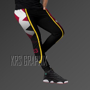 May include: Black athletic pants with a white, burgundy, and black design on the left leg, and a yellow stripe down the side. The number "23" is printed in burgundy. The text "KRS GRAPHIX" is at the bottom. The person is wearing black and white sneakers.