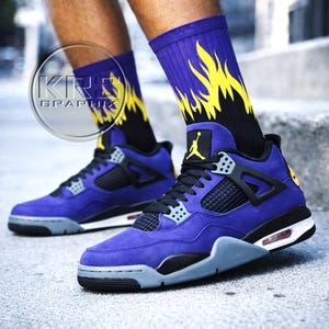 May include: A pair of purple and black Air Jordan 4 sneakers with gray accents and black laces. The sneakers are paired with purple socks featuring a yellow flame design. The Jordan logo is visible in yellow.