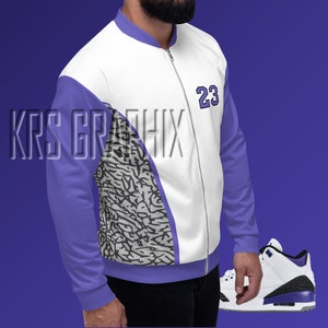 Puede incluir: Chaqueta bomber blanca y morada con cierre de cremallera y el número 23 en el pecho. La chaqueta tiene un diseño estampado gris y blanco en el lado izquierdo. También se muestra una zapatilla blanca y morada.