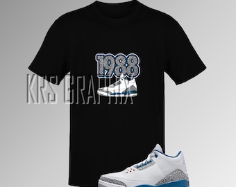 Peut inclure: Un t-shirt noir avec un imprimé de baskets blanches et bleues et le texte "1988" sur le devant. Une basket blanche et bleue avec un motif gris et bleu est également représentée.