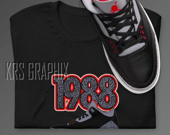 Peut inclure: T-shirt noir avec un motif gris et noir du nombre 1988 avec une chaussure grise et noire sur le dessus du nombre. La chaussure a un accent rouge à l'arrière.