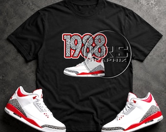 Peut inclure: Un t-shirt noir à manches courtes arborant le nombre 1988 avec un motif imprimé éléphant gris et des contours rouges, placé au-dessus d'une basket blanche, rouge et grise. Le texte RS GRAPHIX est visible dans une superposition circulaire.