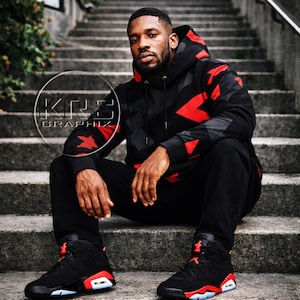 Pu&ograve; includere: Una persona indossa una felpa con cappuccio nera e rossa a fantasia e pantaloni abbinati, seduta su gradini di cemento. La felpa con cappuccio e i pantaloni presentano un design geometrico con dettagli rossi. Le scarpe da ginnastica nere con dettagli rossi e bianchi completano l'outfit. Il testo "KRS Graphix" &egrave; incluso.