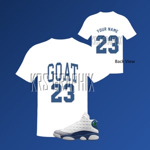 May include: White t-shirt with the word "GOAT" and the number "23" printed in blue. A white t-shirt with the number "23" printed in blue. White and blue basketball shoes.