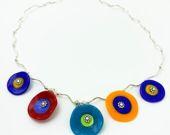 May include: A silver chain necklace with five colorful glass circles. Each circle has a silver center with a small flower design. The circles are red, blue, yellow, blue, and turquoise.