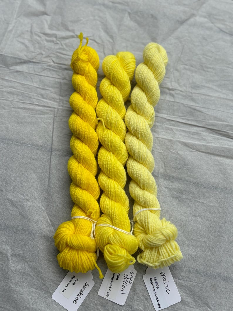 Pu&ograve; includere: Tre gomitoli di filato tinto a mano in tonalit&agrave; di giallo. I gomitoli sono etichettati "Mais", "Giallo morbido" e "Sole".