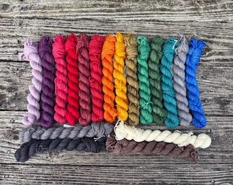 Puede incluir: Una colección de madejas de hilo coloridas en varios tonos, incluyendo morado, rojo, naranja, verde y azul. El hilo está dispuesto sobre una superficie de madera envejecida, mostrando una gama de colores para proyectos de manualidades.