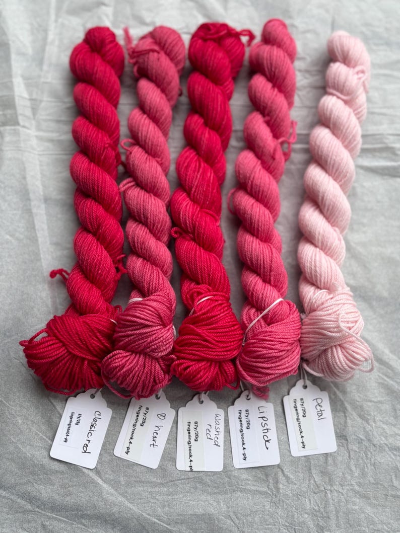 Pu&ograve; includere: Sei gomitoli di filato per calze tinto a mano in tonalit&agrave; di rosa e rosso. I gomitoli sono etichettati con i nomi "Petal", "Lipstick", "Washed Red", "Heart" e "Classic Red".