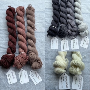Pu&ograve; includere: Assortimento di gomitoli di filato fingering tinto a mano in varie tonalit&agrave; di marrone, grigio e bianco. I gomitoli sono etichettati con i loro nomi e pesi, tra cui "Chocolate", "Bark", "Teddy Bear", "Silver", "Slate", "Charcoal", "Black", "Creamy Natural Sparkle" e "Creamy Natural".