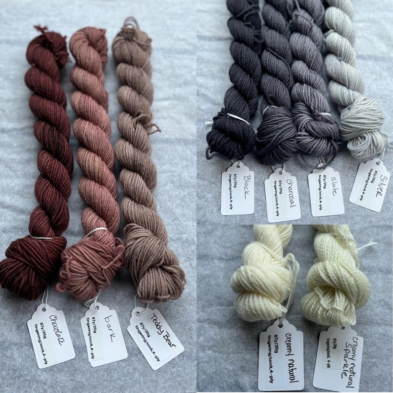 Pu&ograve; includere: Assortimento di gomitoli di filato fingering tinto a mano in varie tonalit&agrave; di marrone, grigio e bianco. I gomitoli sono etichettati con i loro nomi e pesi, tra cui "Chocolate", "Bark", "Teddy Bear", "Silver", "Slate", "Charcoal", "Black", "Creamy Natural Sparkle" e "Creamy Natural".