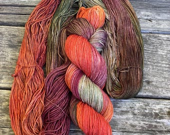 May include: A skein of hand-dyed yarn featuring a variegated color palette of deep burgundy, burnt orange, olive green, and earthy brown tones. The yarn is twisted into a thick, textured bundle, showcasing a blend of warm and muted autumn-inspired hues.