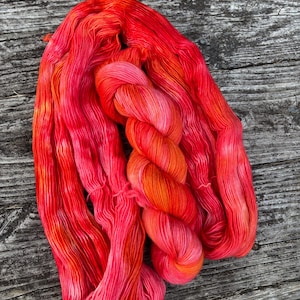 May include: A skein of hand-dyed yarn in vibrant shades of red, orange, and pink. The yarn is arranged in a loose, flowing pattern, showcasing the color variations. The yarn is likely made of a soft fiber, perfect for knitting or crocheting projects.