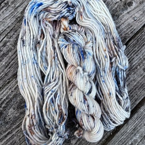 May include: A skein of hand-dyed yarn in shades of white, blue, and brown. The yarn is made of a soft, fluffy fiber and has a speckled appearance.
