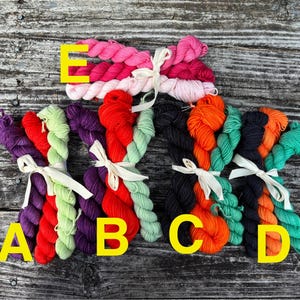 May include: Bundles of colorful yarn in shades of pink, red, purple, green, orange, and black, tied with white ribbons. The yarn is arranged on a weathered wooden surface, with yellow letter labels.